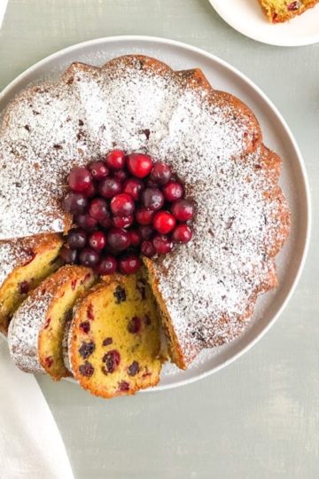 Bundt cake dusted with powdered sugar on large round white place with whole fresh cranberries in the center and a few slices of cake, next to white kitchen towel. One slice on small round white plate and gold tea strainer with powdered sugar in the background.